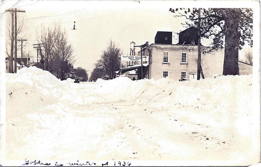 The front of a black and white postcard titled 'Gobles in winter 1936' shows a snowy scene looking down a plowed road. To the left of the road are trees, to the right, a brick building and in the distance a water tower.
